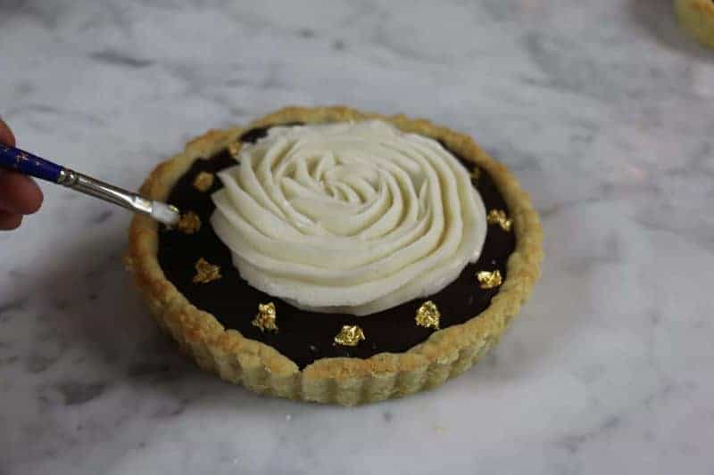 A photo of edible gold leaf carefully being placed on top of a chocolate tart.