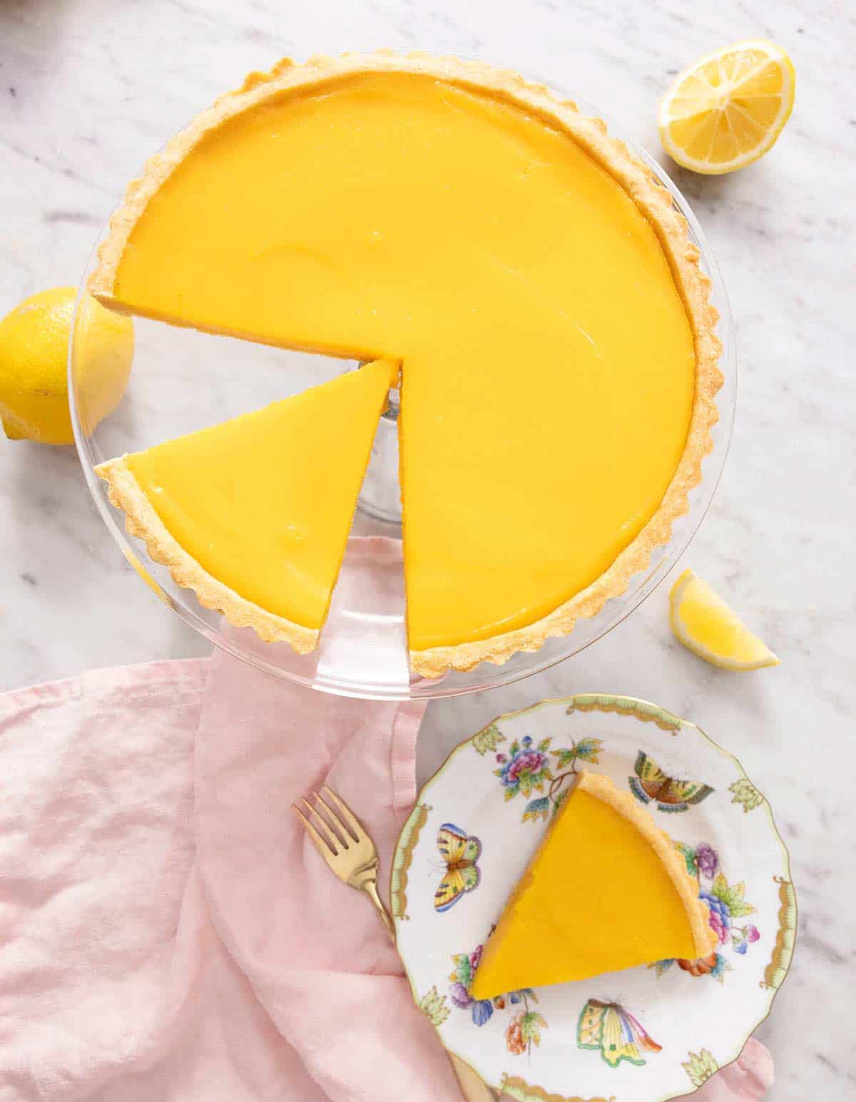 An overhead shot of a lemon tart on a cake stand and a slice on a plate