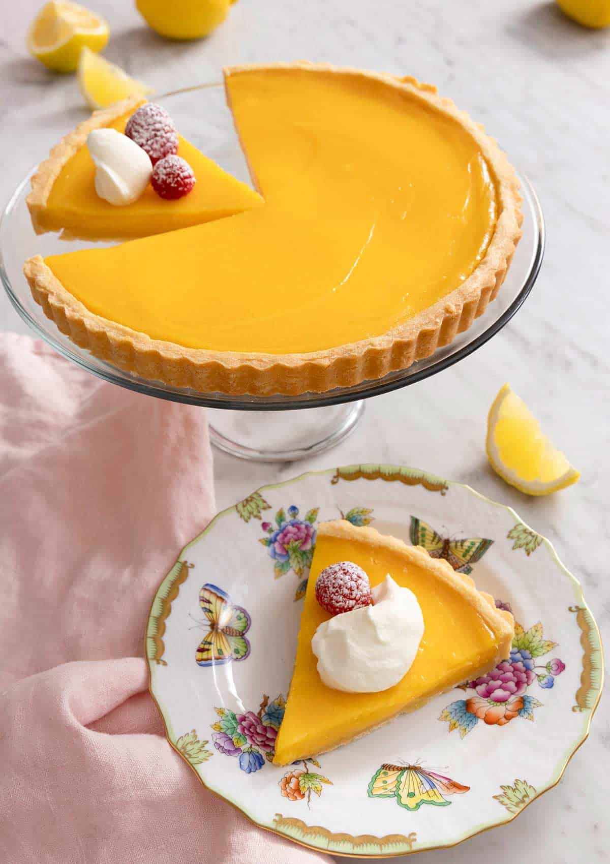 A lemon tart on a cake stand with a slice plated in front.