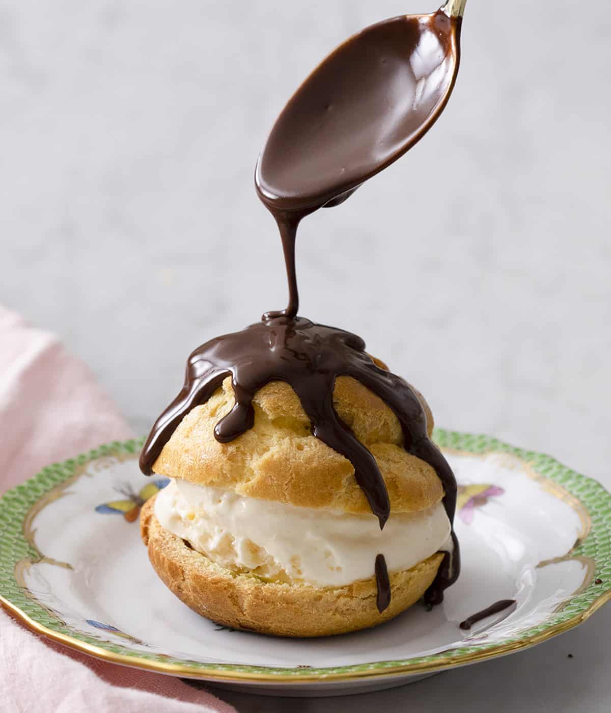 A spoonful of melted chocolate drizzled overtop of a profiterole on a plate.