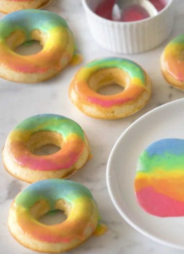 Rainbow donuts on a marble surface with a plate with the glaze beside it.