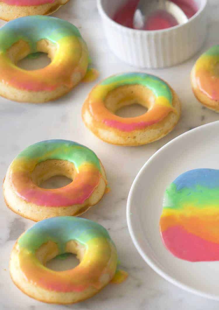 Rainbow donuts on a marble surface with a plate with the glaze beside it.
