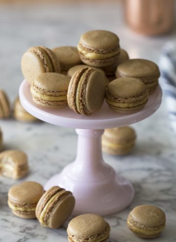 Pumpkin macarons piled onto a cake plate with others surrounding it.
