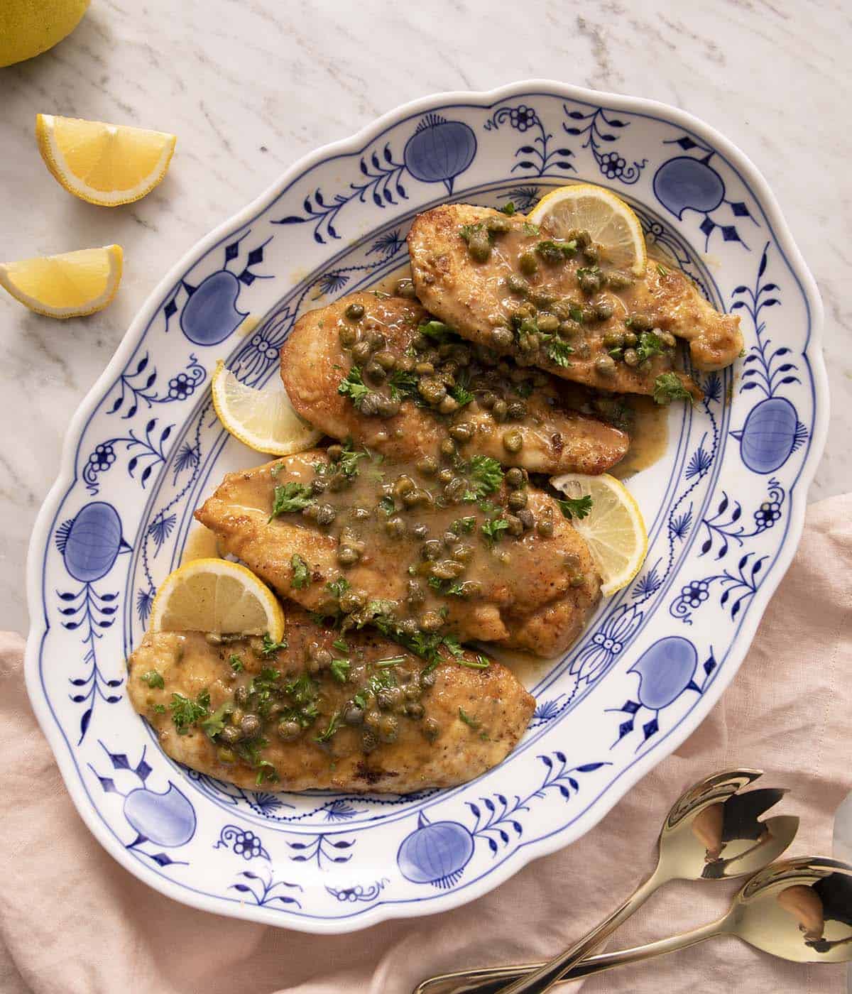 Overhead view of an oval platter of chicken piccata with lemon slices as garnish.