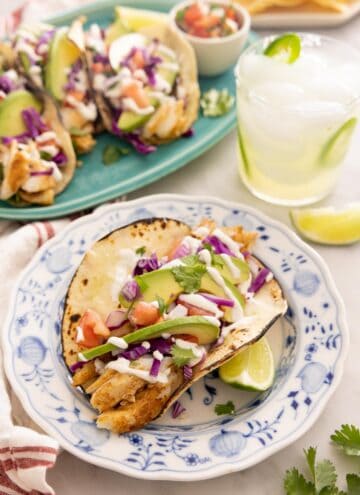 A freshly made fish taco on a blue and white plate in front of a platter of fish tacos.