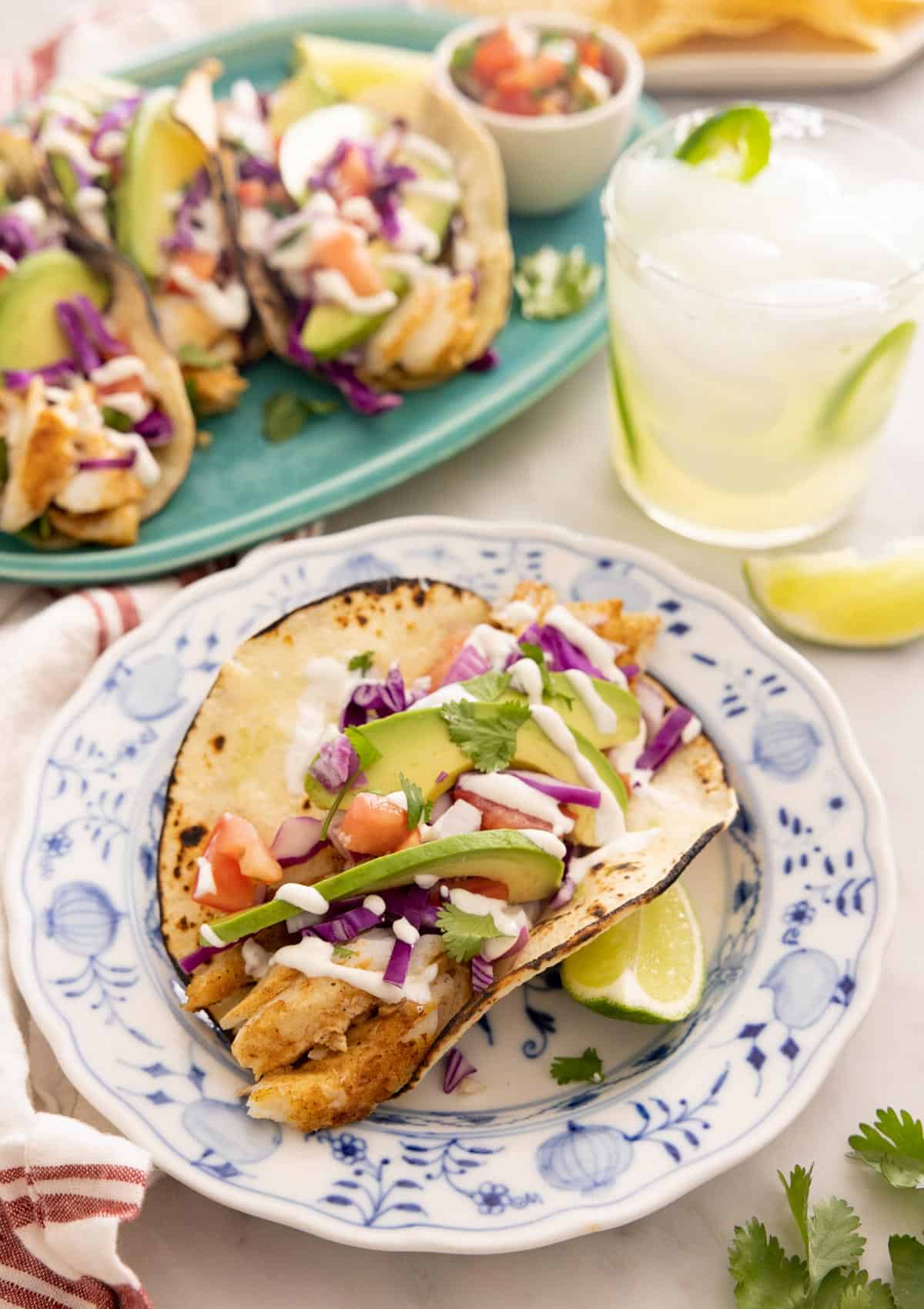 A freshly made fish taco on a blue and white plate in front of a platter of fish tacos.