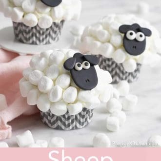 Pinterest graphic of three sheep cupcakes with one on a mini cake stand. Multiple mini marshmallows scattered around with a pink napkin.