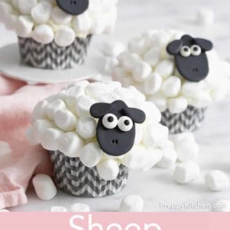 Pinterest graphic of three sheep cupcakes with one on a mini cake stand. Multiple mini marshmallows scattered around with a pink napkin.