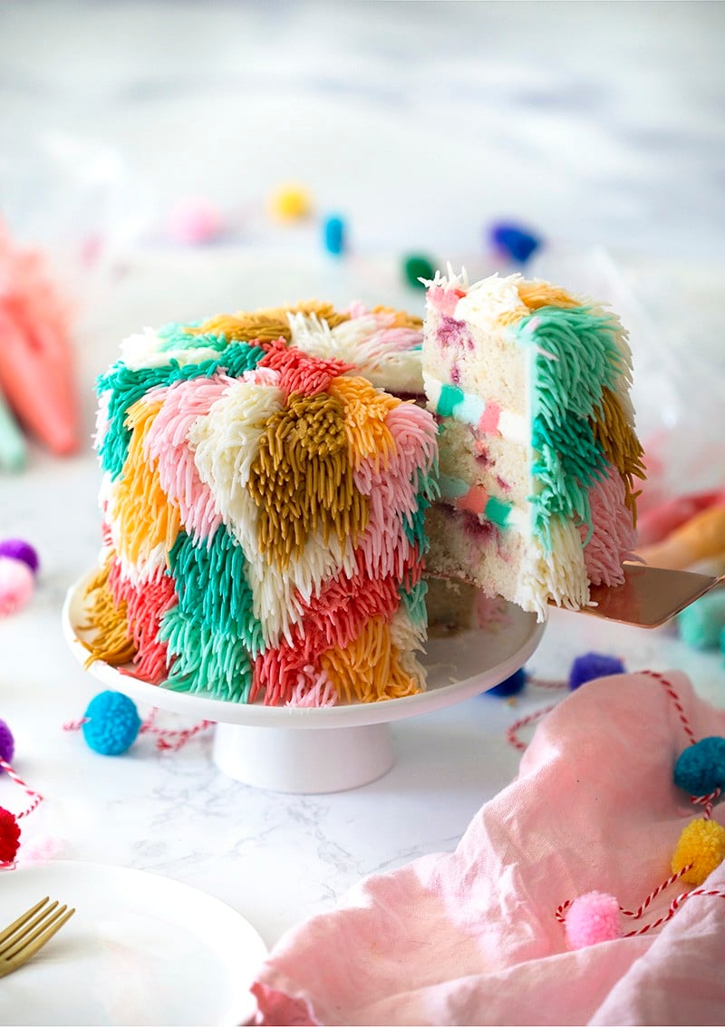 Photo of a colorful shag cake on a white cake stand with a piece being removed