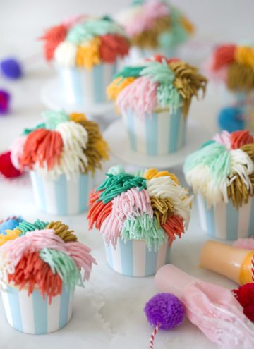Multiple cute cupcakes with multi-colored frosting. Mini pom-poms and frosting bags scattered around.