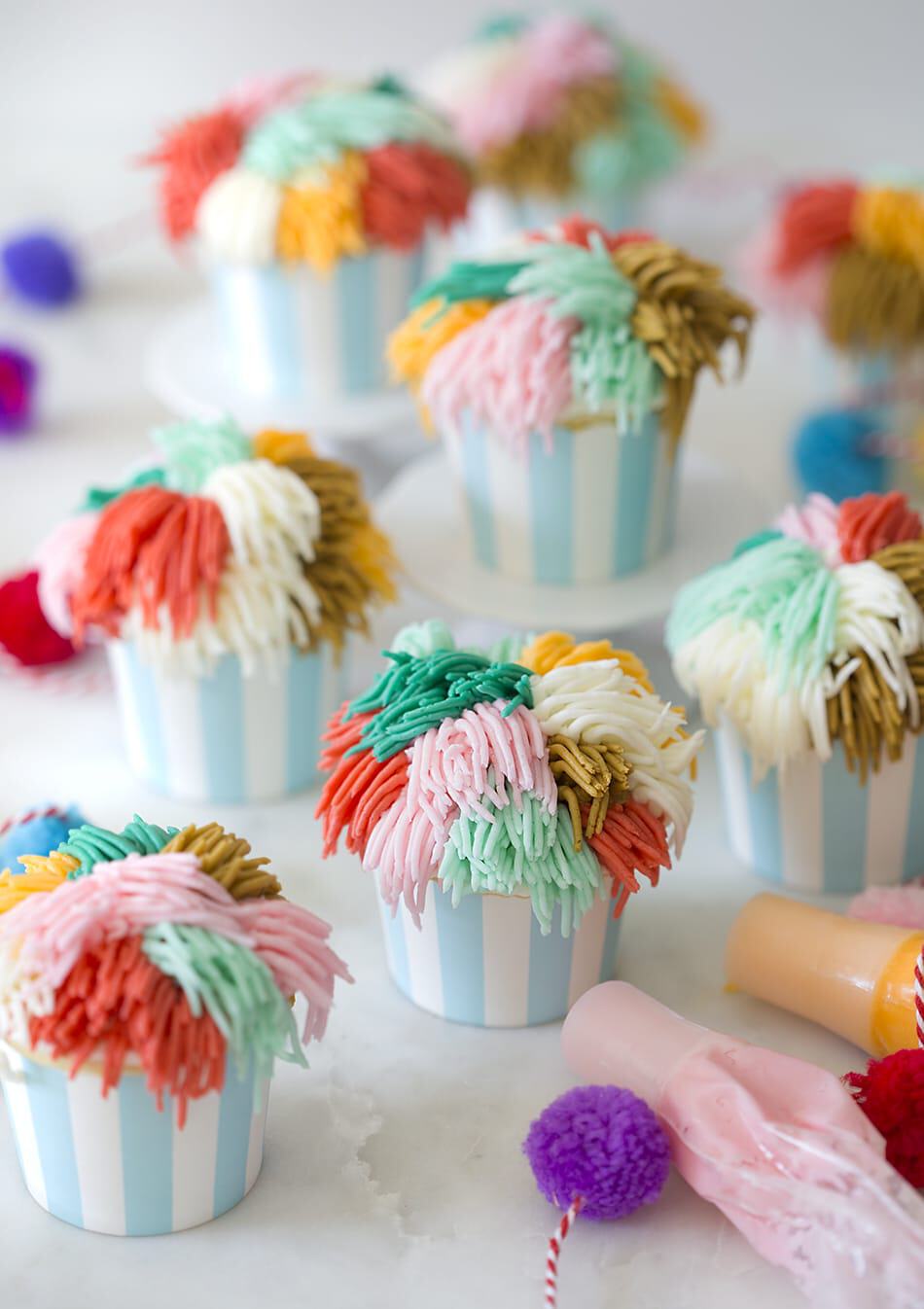 Multiple cute cupcakes with multi-colored frosting. Mini pom-poms and frosting bags scattered around.