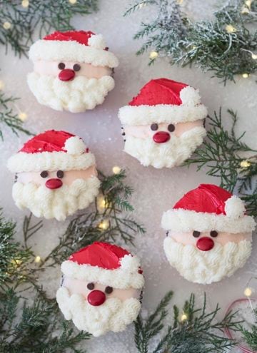 A photo showing a group of santa cupcakes on a white marble table with Christmas lights and pine needles.