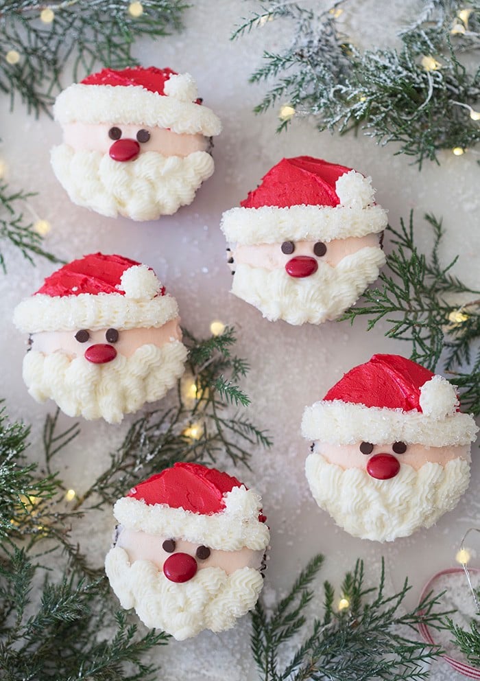 A photo showing a group of santa cupcakes on a white marble table with Christmas lights and pine needles.