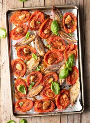 Overhead view of roasted tomatoes with shallots, garlic, and basil on a sheet pan.