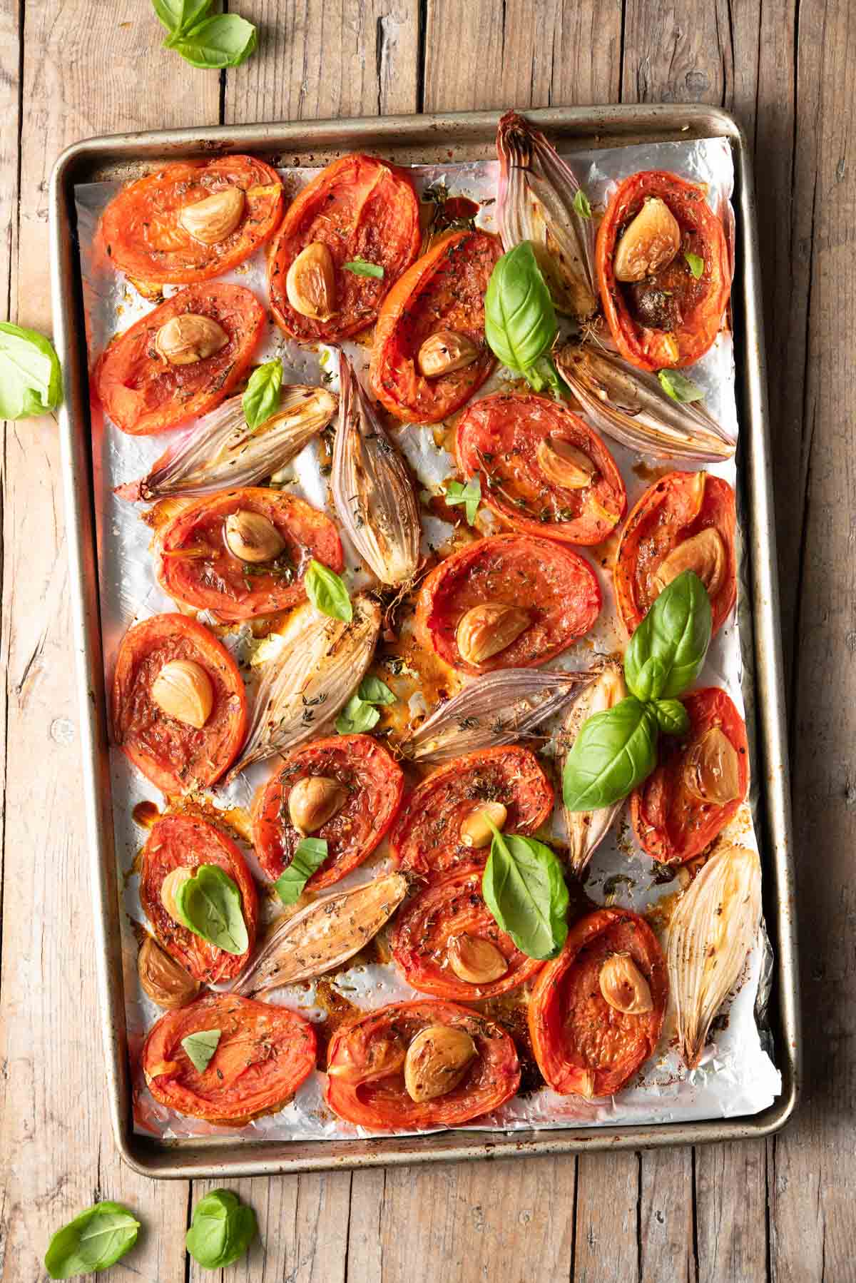 Overhead view of roasted tomatoes with shallots, garlic, and basil on a sheet pan.