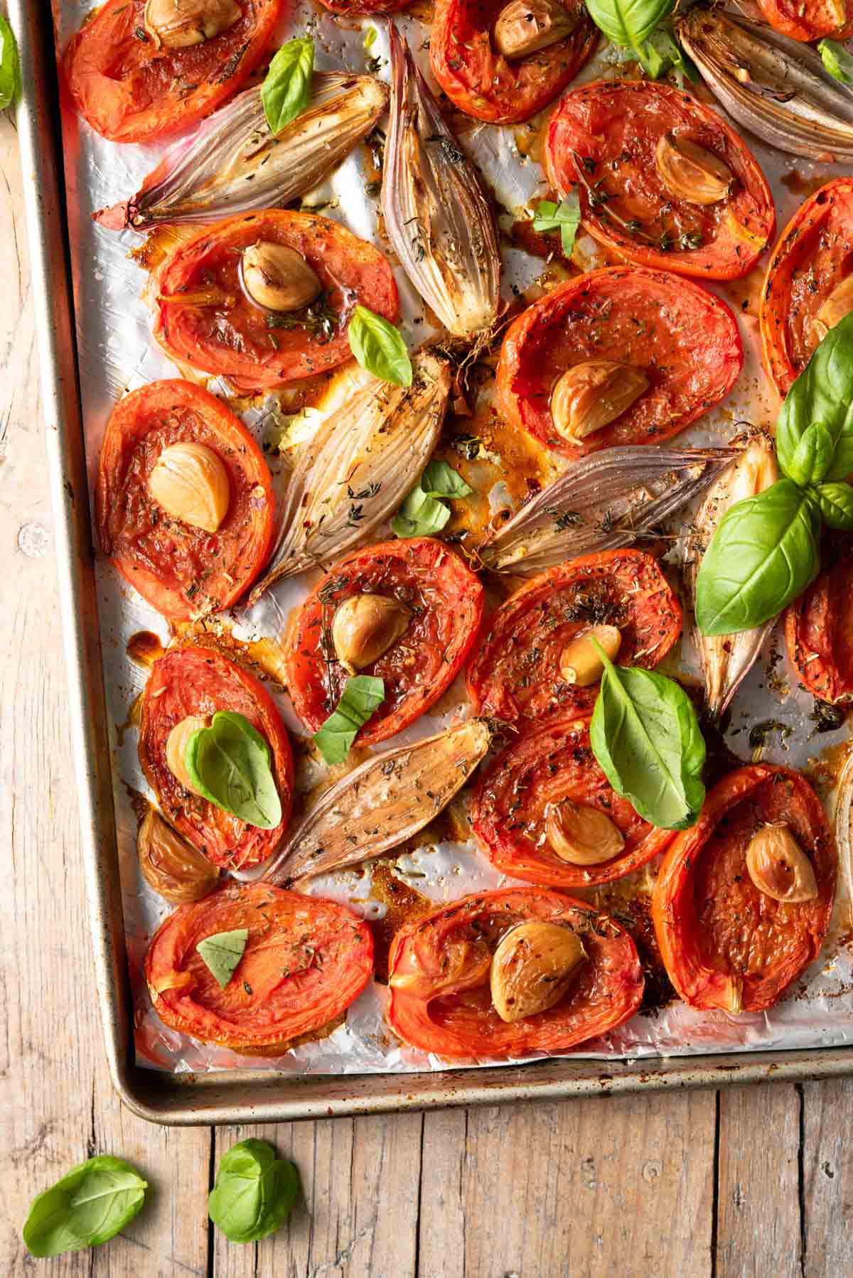 A close up of roasted tomatoes, garlic and shallots on a baking tray