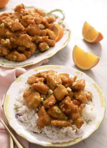 Rice and orange chicken on a plate with orange wedges in the background