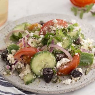 Greek salad on a spekled white plate next to a glass on white wine.