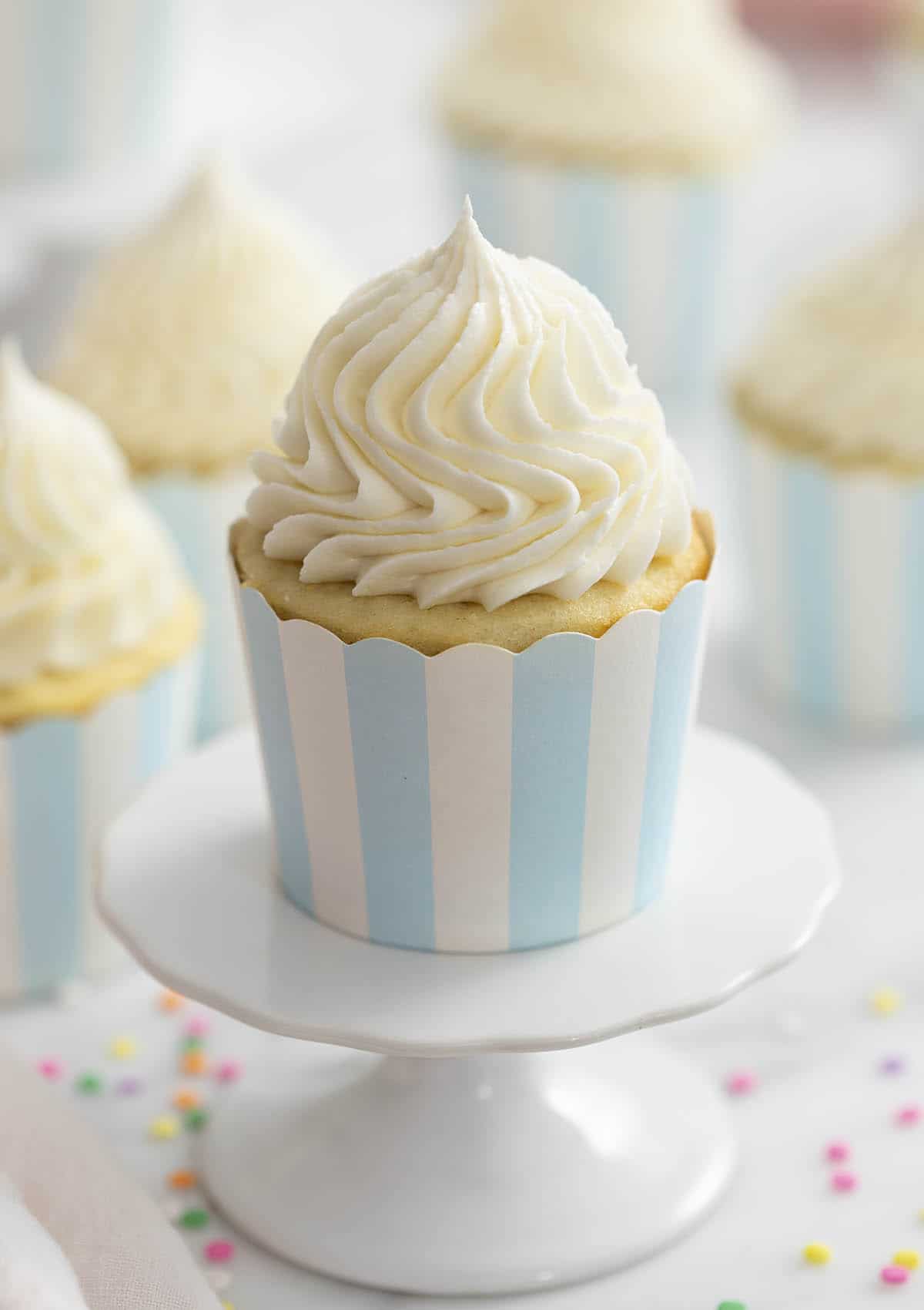 A photo of a vanilla cupcake on a cupcake stand with vanilla buttercream frosting beautifully piped on top.