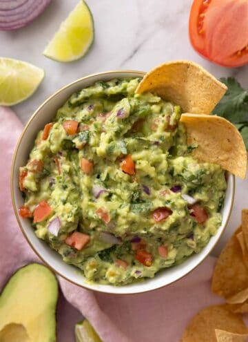 An overhead shot of a bowl of guacamole with tortilla chips inside of the bowl and ingredients scattered around the side.