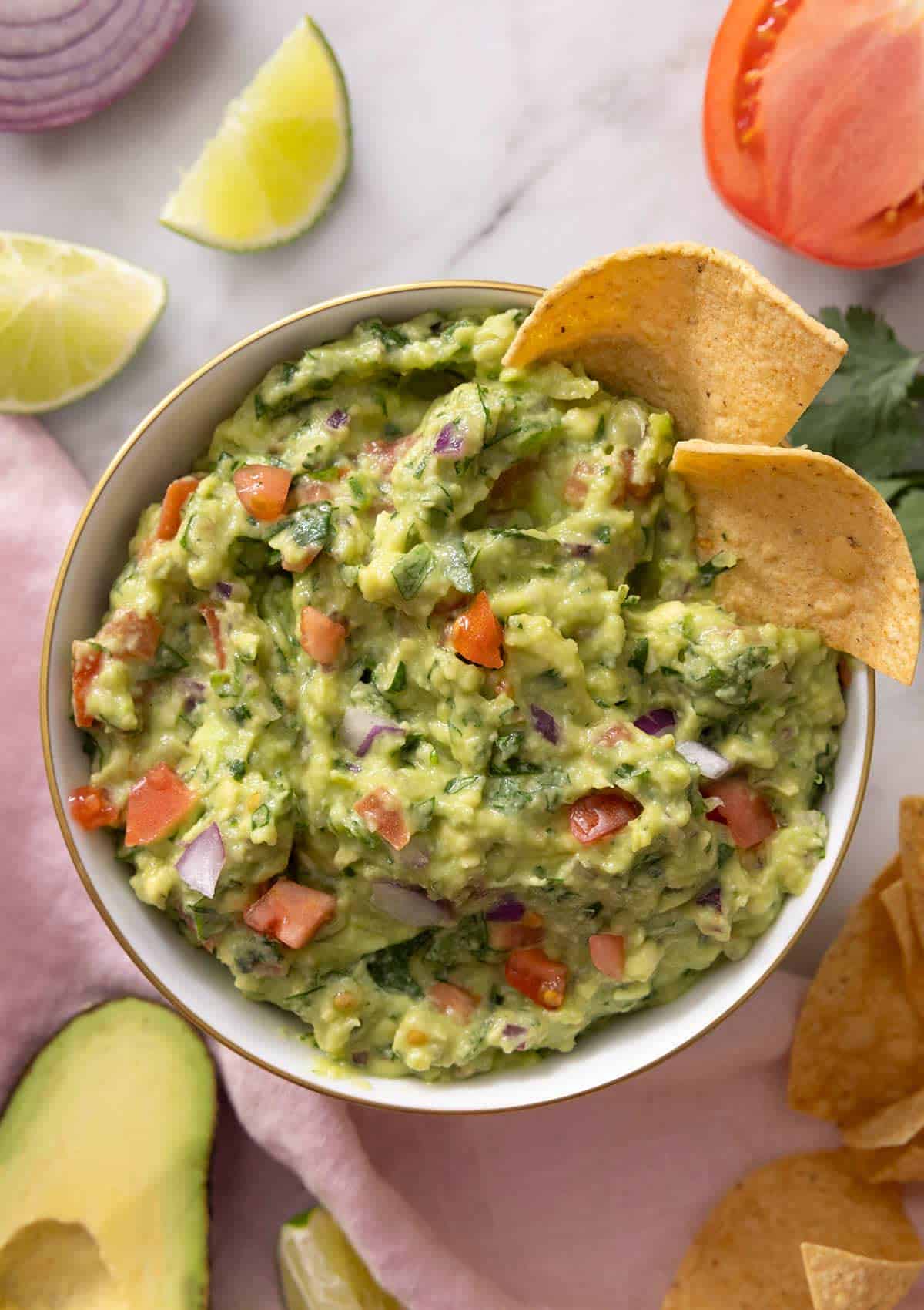 An overhead shot of a bowl of guacamole with tortilla chips inside of the bowl and ingredients scattered around the side.