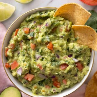 Pinterest graphic of a bowl of guacamole with tortilla chips. Guacamole ingredients scattered around the marble surface.