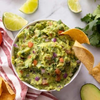 Pinterest graphic of an overhead view of a bowl of guacamole with tortilla chips. Guacamole ingredients scattered around the marble surface.