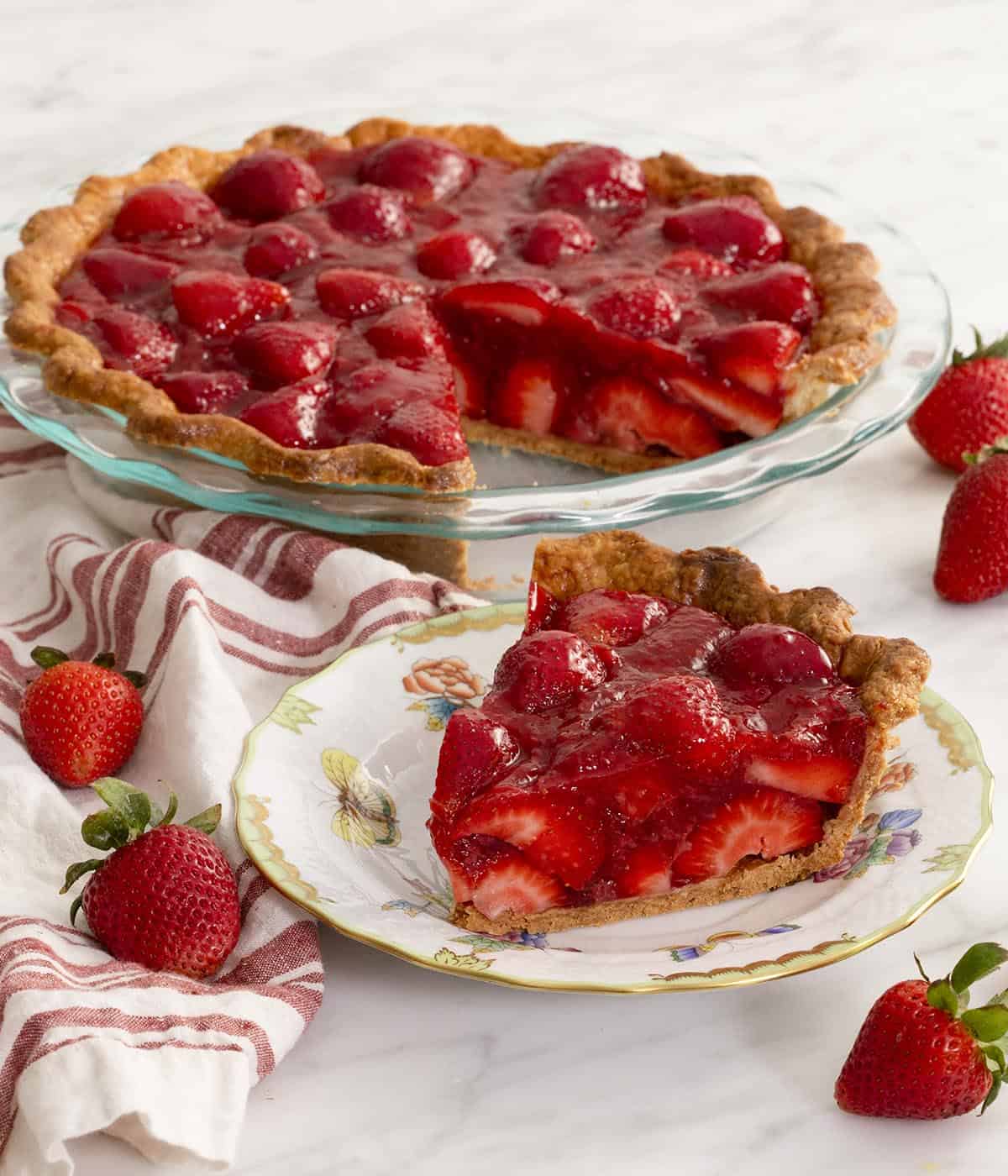 A fresh strawberry pie with a slice in the foreground on a plate on a white marble table. Fresh strawberries scattered around.