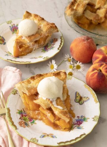 Two pieces of peach pie on porcelain plates with ice cream on top.