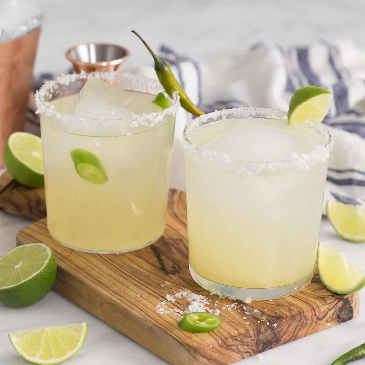 Two margarita cocktails on the rocks with salt in a tumbler. Lime wedges scattered around.