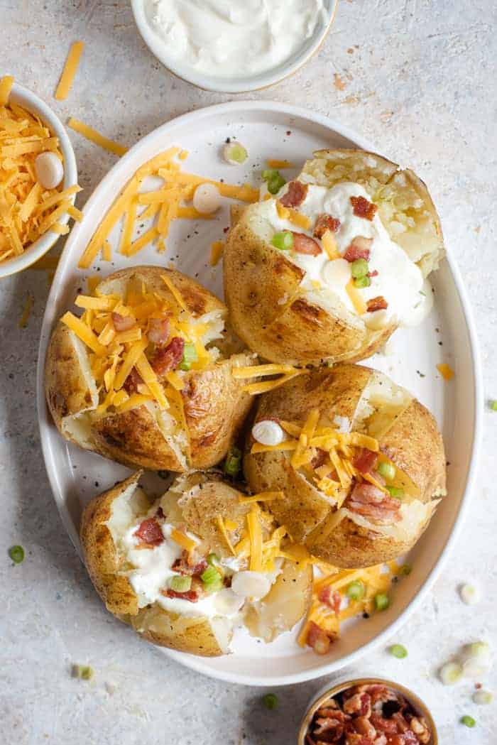An overhead shot of instant pot baked potatoes on a serving plate with an assortment of toppings. More toppings in bowls off to the side.