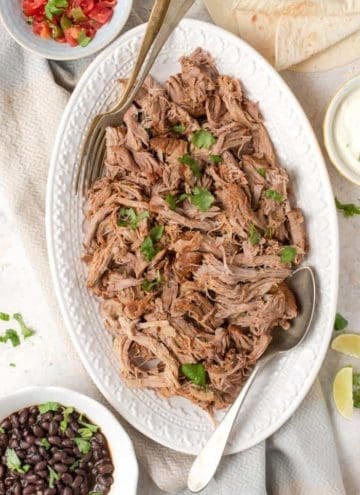 An overhead shot of instant pot pulled pork on a white platter. A bowl of black beans and salsa on the side.