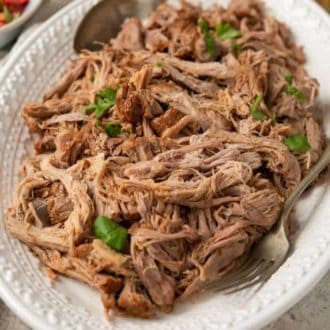 Angled view of a platter of Instant Pot pulled pork with a spoon and fork. Topped with chopped parsley as garnish.