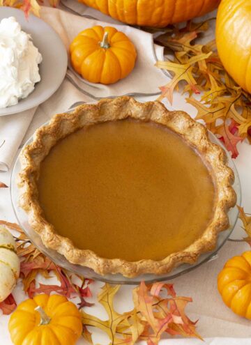 A pumpkin pie next to lots of mini pumpkins and a bowl of whipped cream.