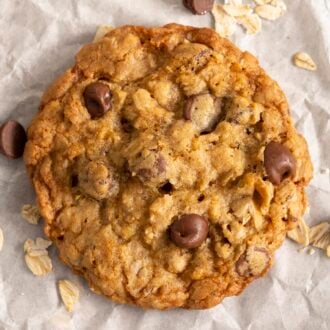 An oatmeal chocolate chip cookie next to some oats and chocolate chips on a piece of parchment paper.