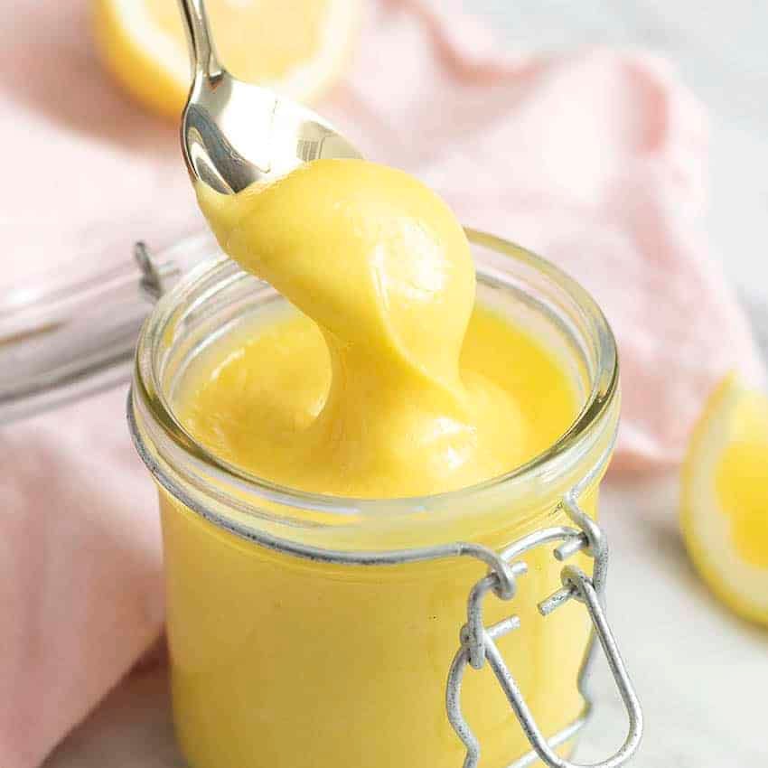 Thick homemade lemon curd getting spooned out of a glass jar.