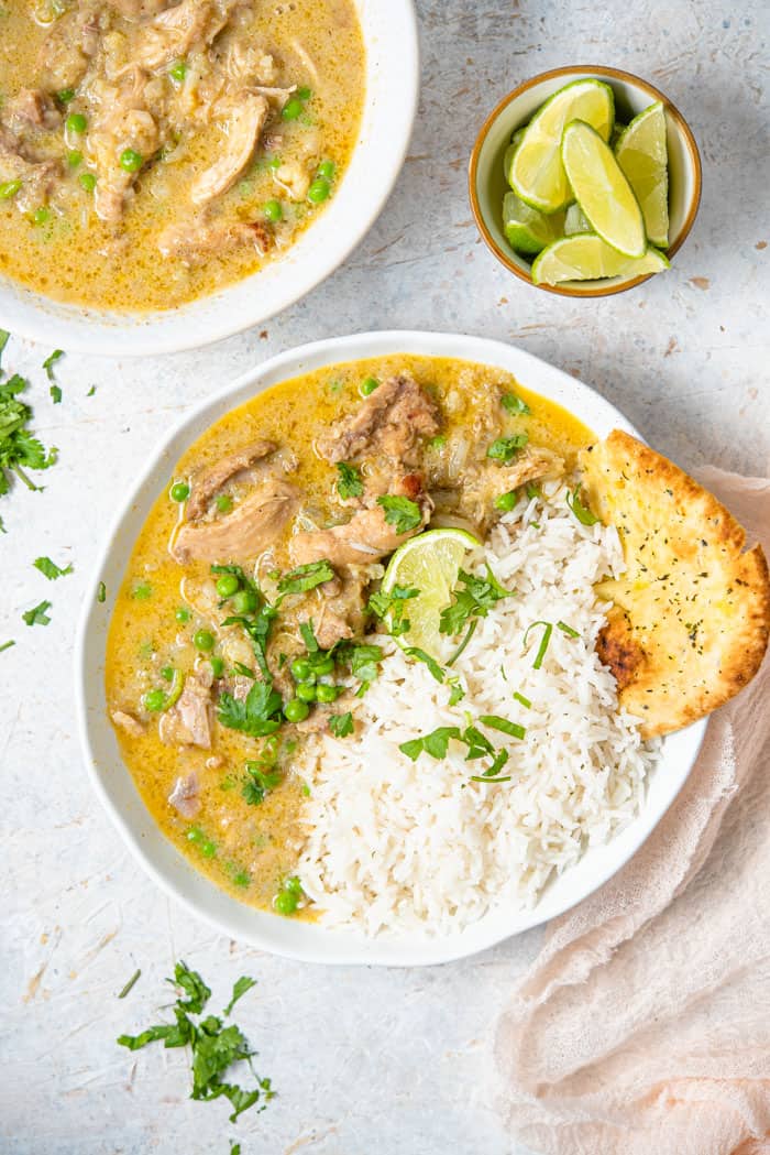 An overhead shot of chicken curry in a bowl with rice and naan. A bowl of lime wedges and just curry on the side.