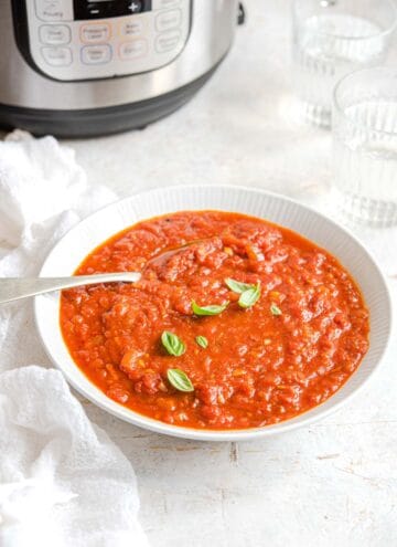 A white bowl of Instant Pot spaghetti sauce topped with some basil leaves with a spoon inside. Instant Pot in the back with two glasses of water.