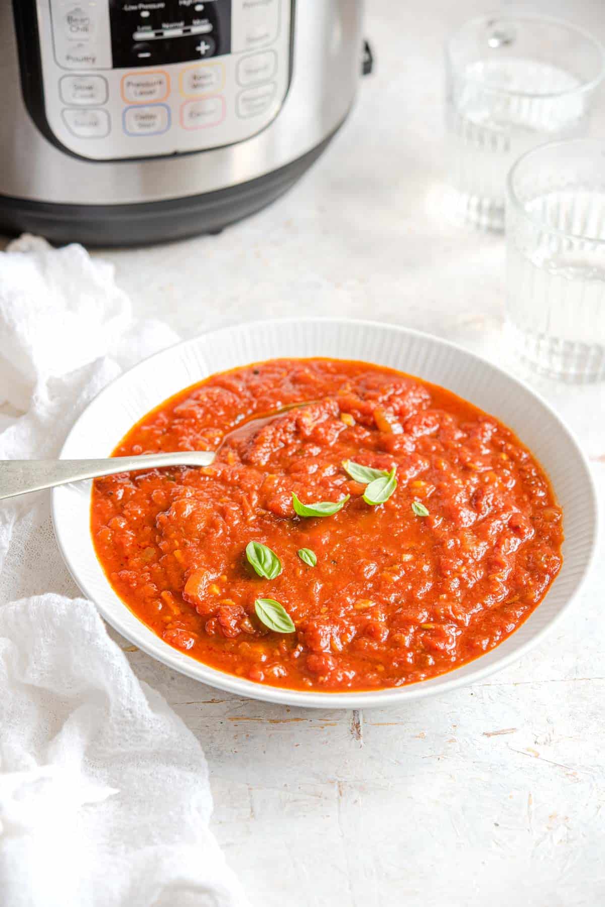 A white bowl of Instant Pot spaghetti sauce topped with some basil leaves with a spoon inside. Instant Pot in the back with two glasses of water.