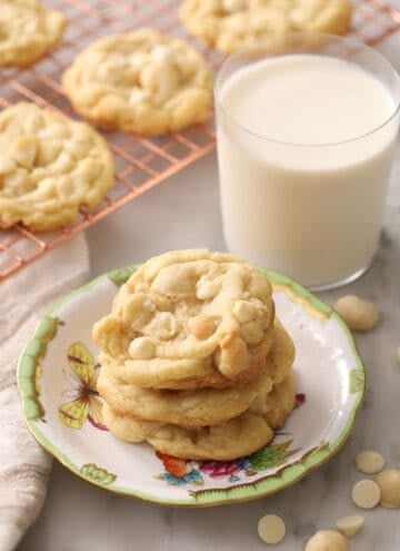 Three white chocolate macadamia nut cookies stacked on a plate alongside a glass of milk.
