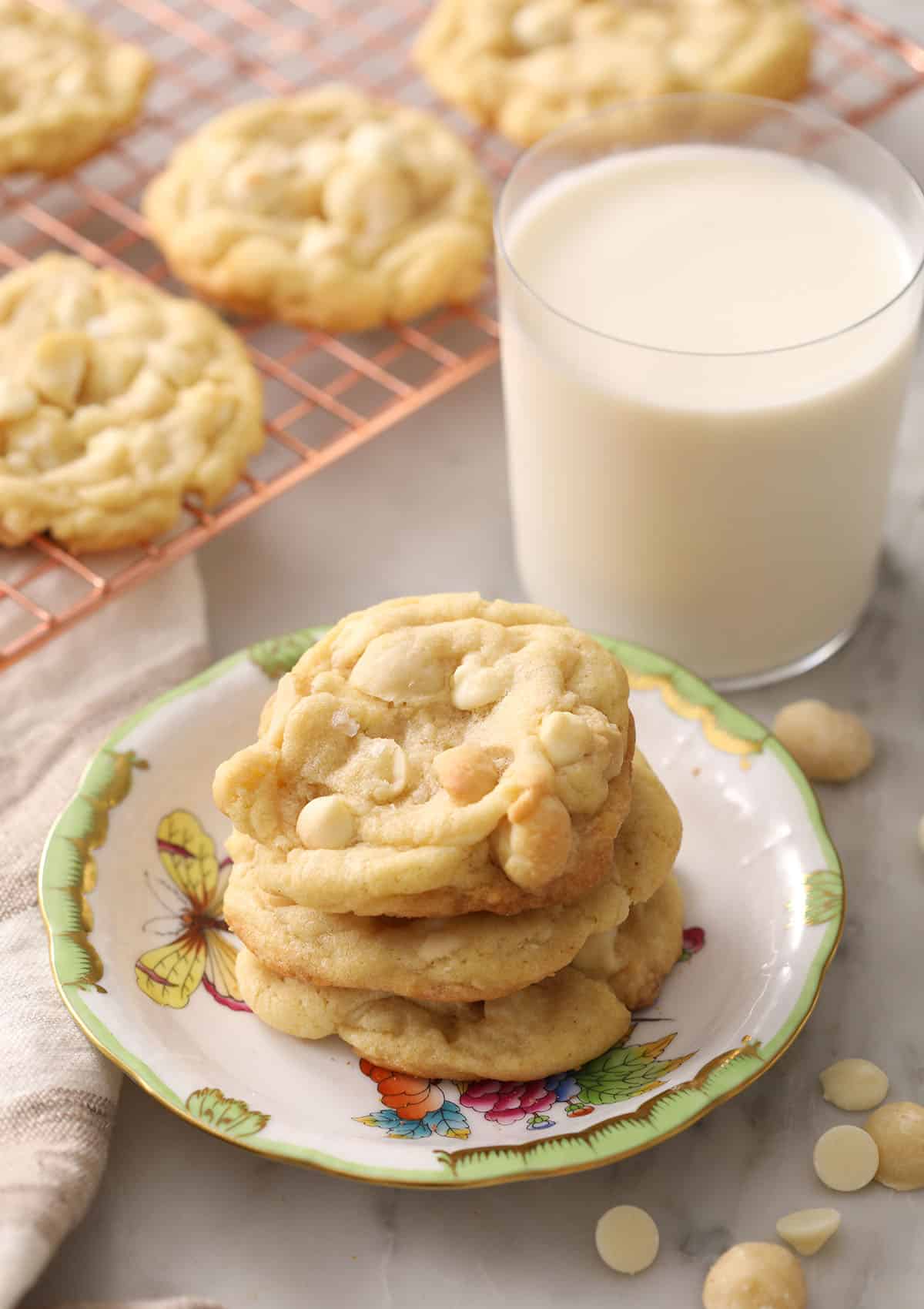 Three white chocolate macadamia nut cookies stacked on a plate alongside a glass of milk.