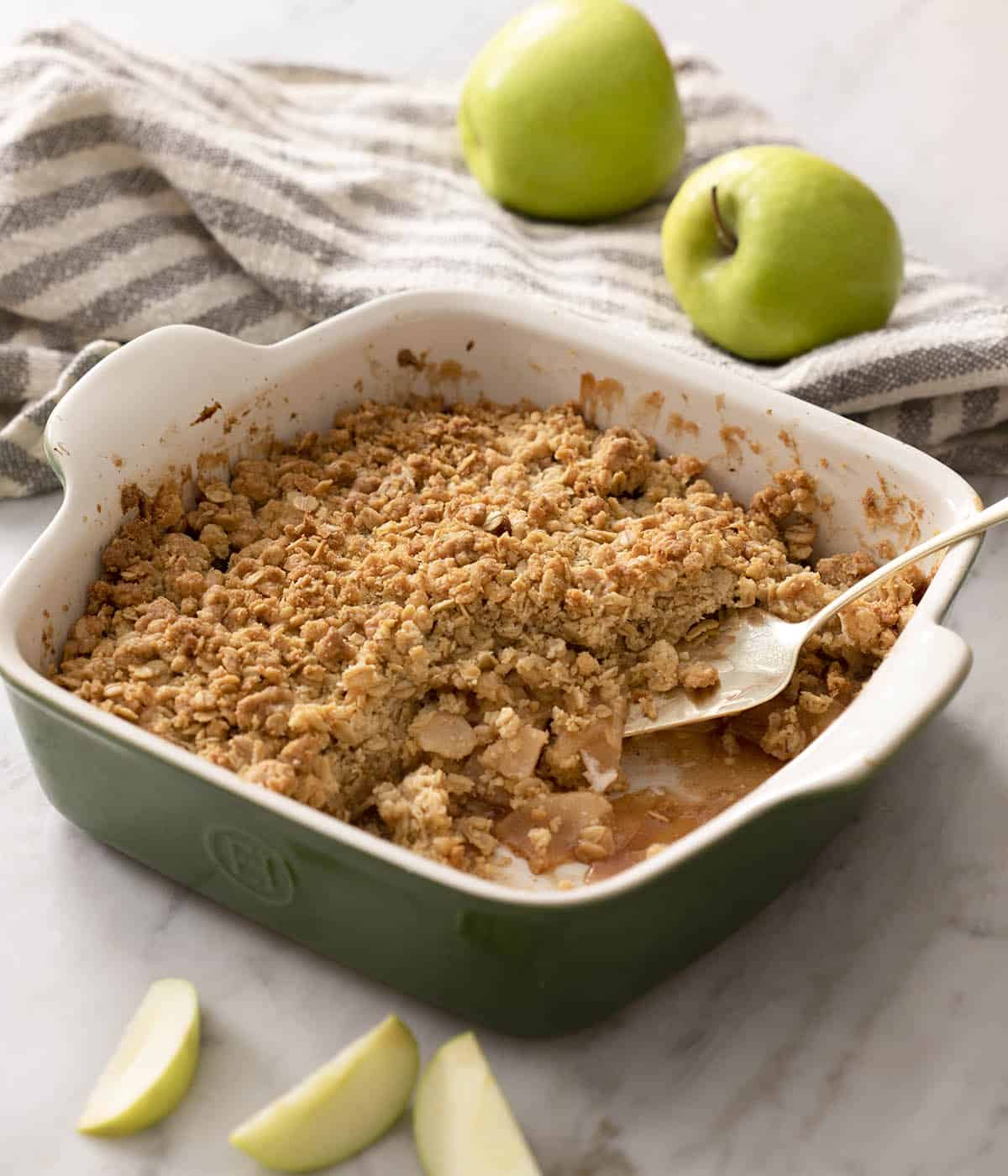 A baking dish of apple crisp on a marble counter with a serving spoon tucked into the crisp. Apples in the background.