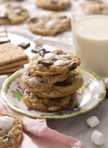 A stack of three s'more cookies on a plate with a glass of milk in the back.