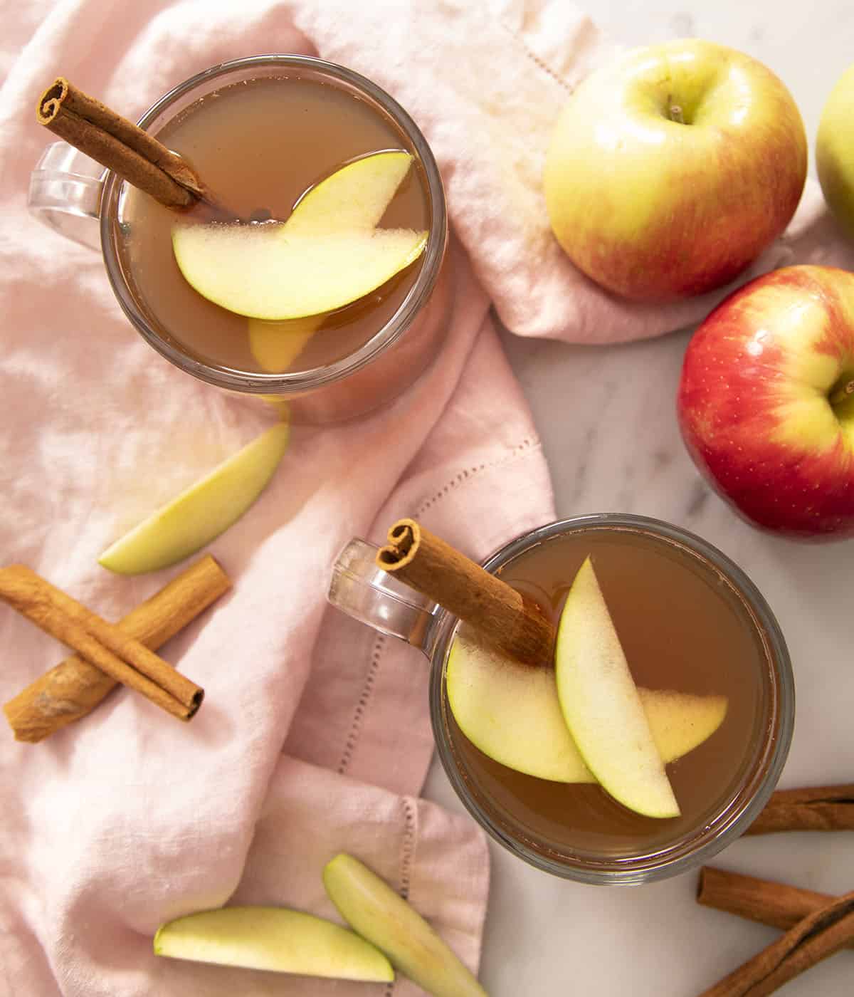 Two glasses of apple cider garnished with cinnamon sticks and apple slices on a marble counter..