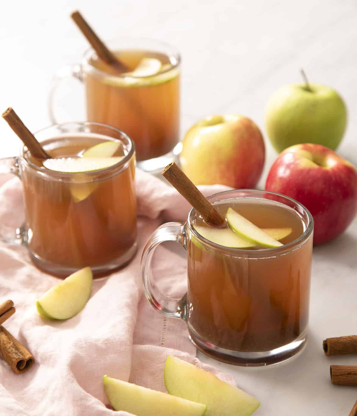 Three glasses of apple cider garnished with cinnamon sticks and apple slices.
