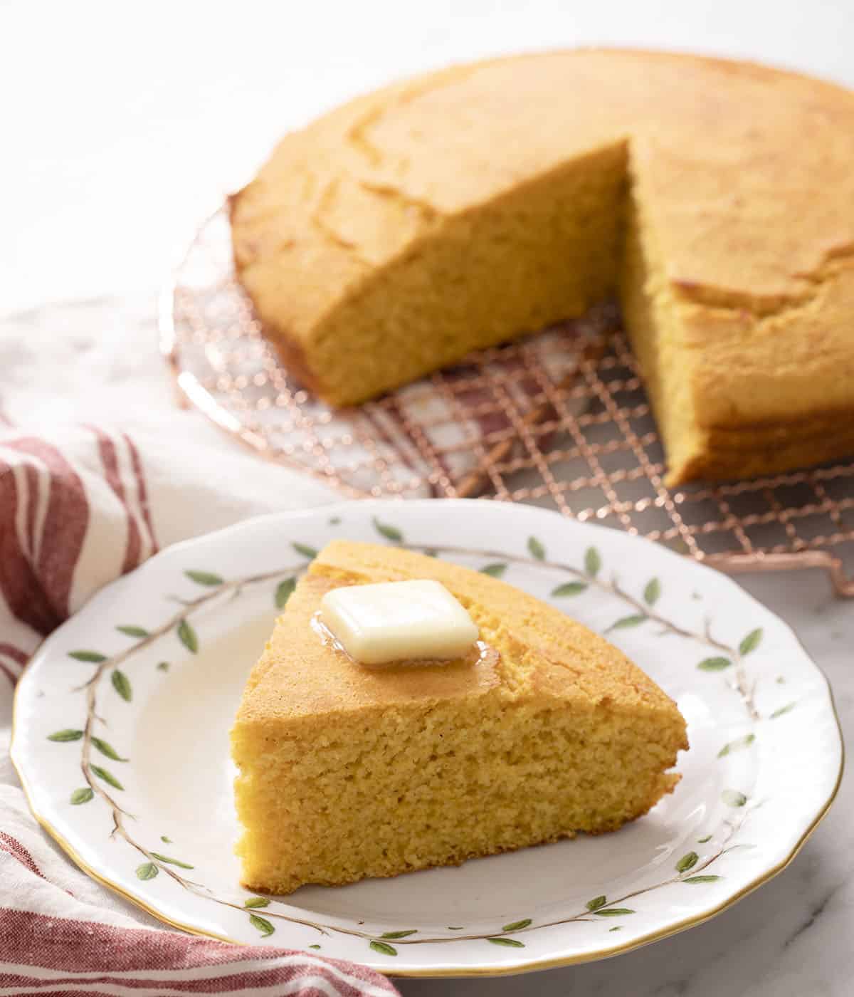 A piece of cornbread on a plate in front of a cornbread loaf on a wire rack.