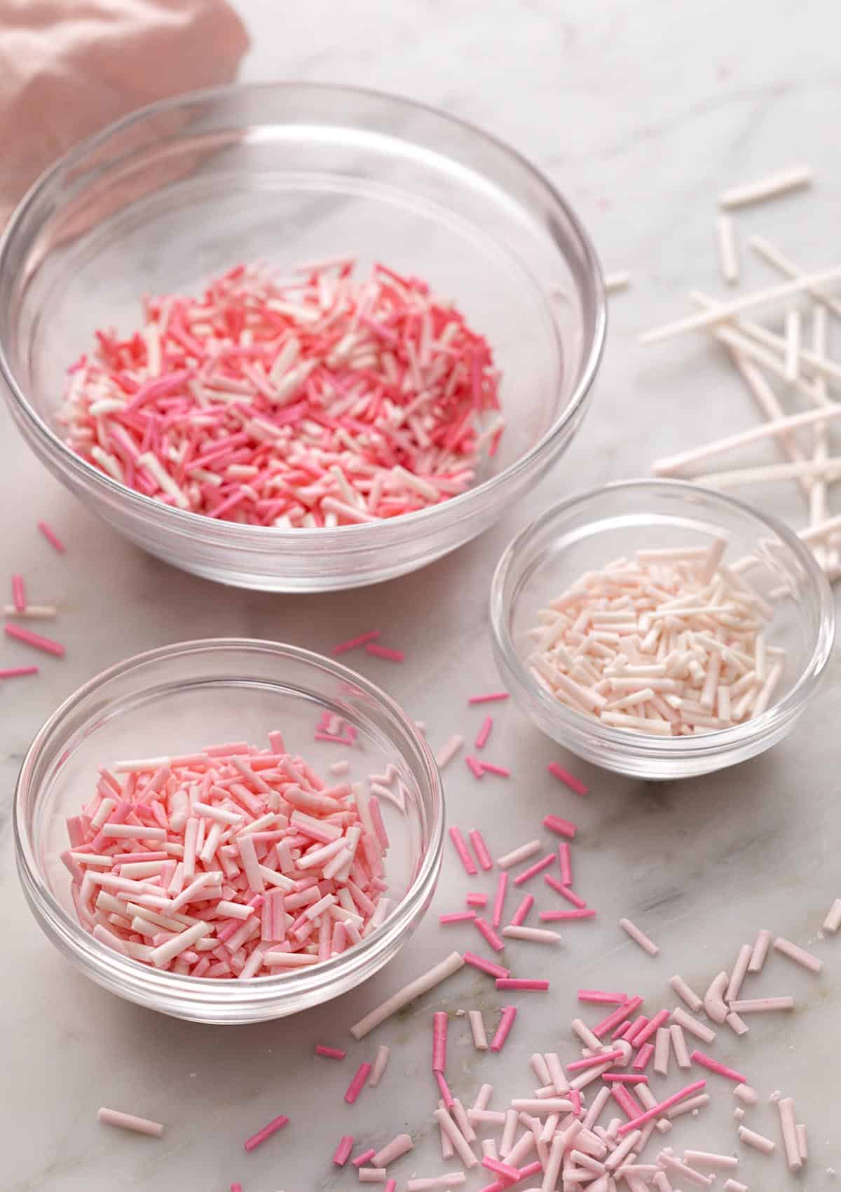 Pink homemade sprinkles in glass bowls on a marble counter.
