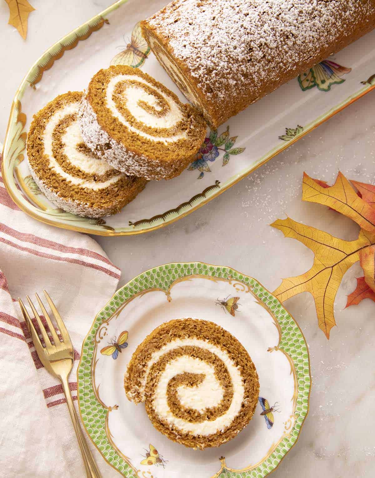 A piece of a pumpkin roll cake on a green and white plate with the rest of the half-sliced cake in the background. 