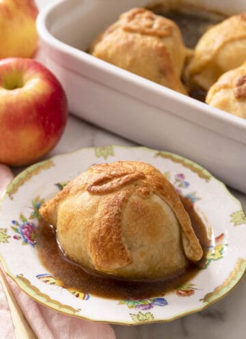 An apple dumpling on a porcelain plate. More apple dumplings in a baking dish in the background.