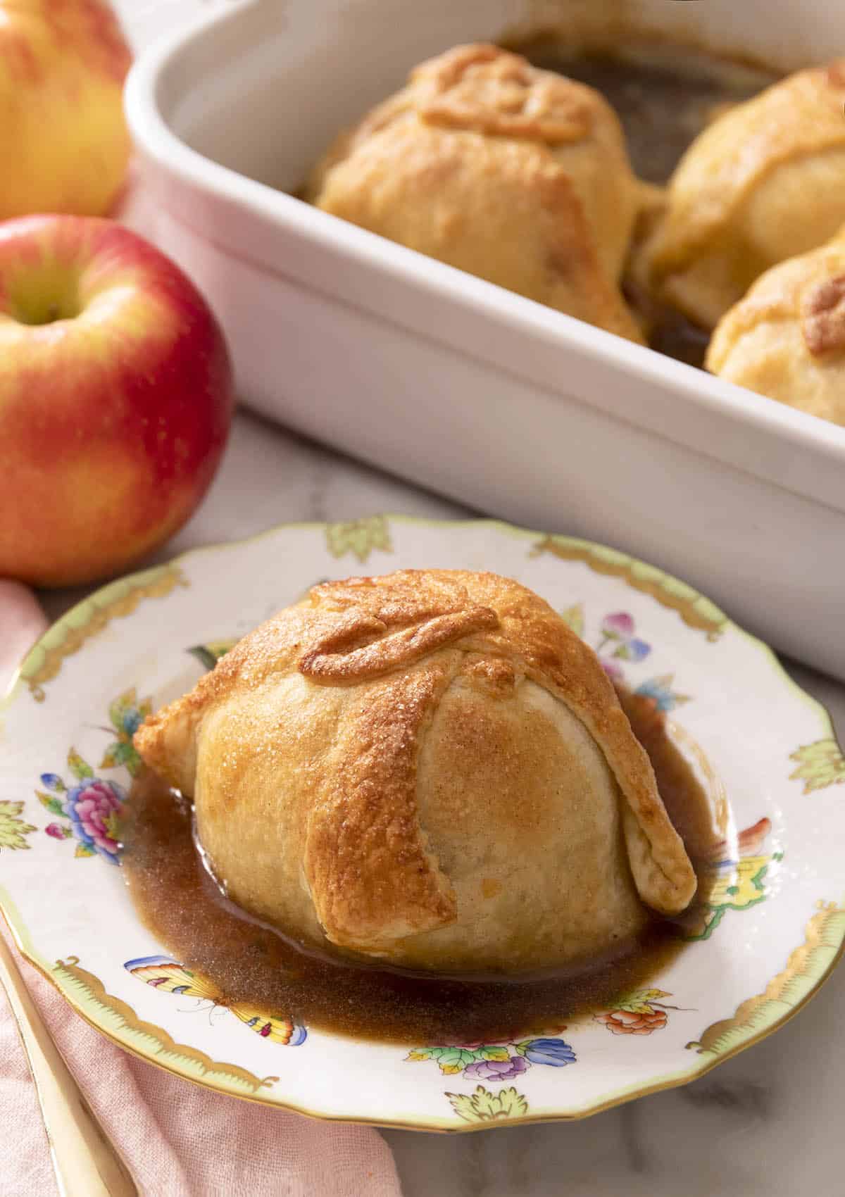 An apple dumpling on a porcelain plate. More apple dumplings in a baking dish in the background.